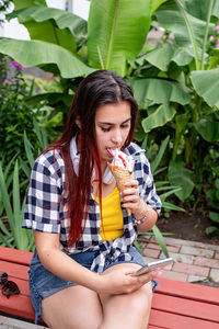 Portrait of smiling young woman eating ice cream sitting against plants