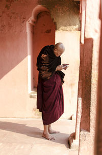 Side view of monk using smart phone while standing against wall