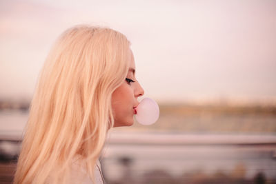 Young woman blowing bubble gum while walking on bridge at sunset