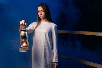 Young woman with illuminated oil lamp standing in forest at dusk