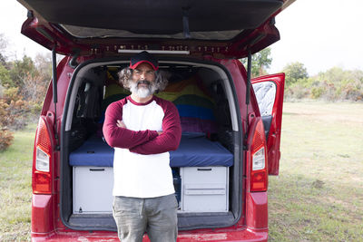 Close-up of a handsome man in a cap standing by a camping van, looking at the camera and smiling.