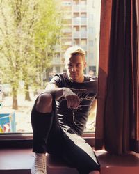 Young man looking at camera while sitting on window