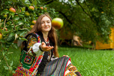 Girl in embroidery sits on the grass under an apple tree