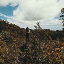 Man standing on rock amidst plants against cloudy sky