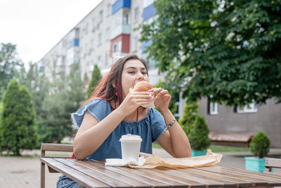 Hungry stylish woman, enjoying eating a burger outdoors, dressed in jeans shirt, wearing sunglasses
