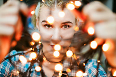 Close-up portrait of young woman holding illuminated string lights at home