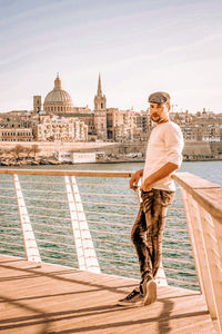 Portrait of young man leaning on railing against river in city