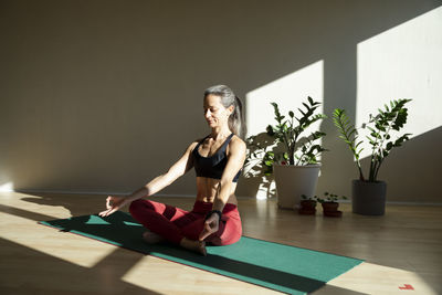 Yogini meditating on exercise mat in living room on sunny day