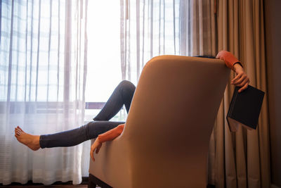 Full length of woman holding book while relaxing on armchair at home