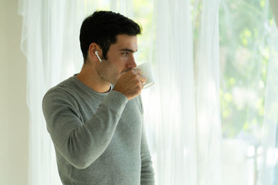 Side view of young man looking away while standing against window