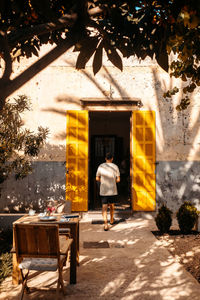 Back view of unrecognizable male tourist in casual clothes walking in backyard towards entrance of house with yellow shutters during summer holidays in balearic islands