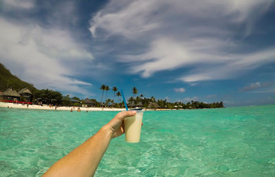 Cropped hand holding drink over sea against sky