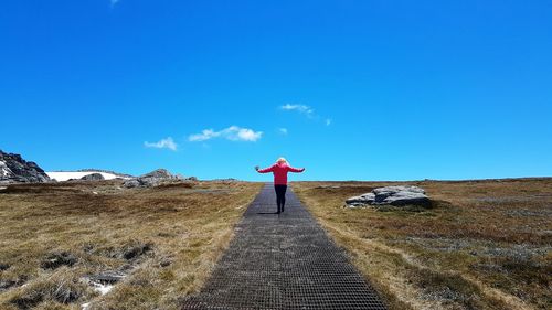 Rear view of woman with arms outstretched standing on field against blue sky during sunny day