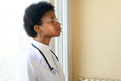 Side view of competent young african american female physician in white medical coat and eyeglasses with stethoscope closed eyes while standing in clinic