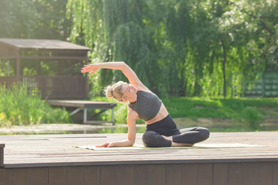 A woman sitting on a wooden platform by a pond and performing a body tilt sideways
