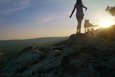 Woman standing with dog on rock against sky during sunset