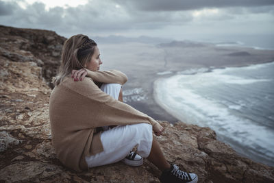 Woman sitting on rock at shore against sky