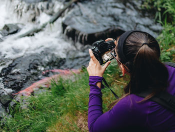 Rear view of woman photographing by stream in forest