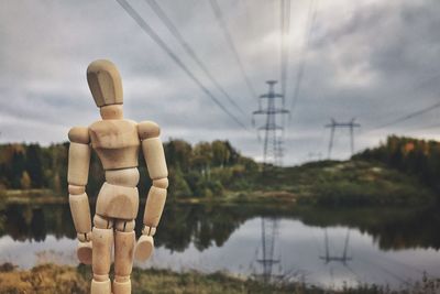 Close-up of wooden figurine against electricity pylon and cloudy sky