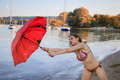 Woman in bikini holding red umbrella while standing at lakeshore