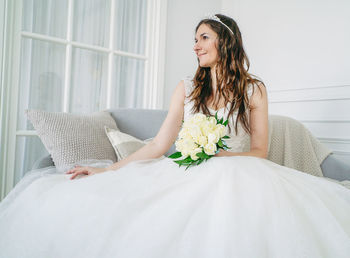Smiling bride holding bouquet while sitting on sofa at home