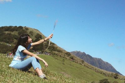 Full length of woman holding grass while sitting on hill against clear blue sky