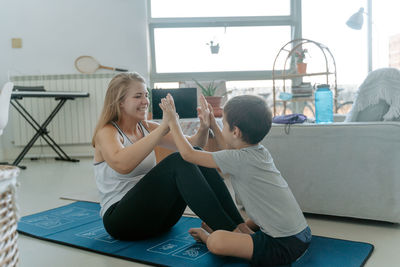 Side view of cheerful young woman resting from doing workout on mat giving son high five at home