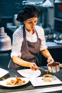 Adult woman in apron and hat preparing and serving dishes on kitchen in modern restaurant