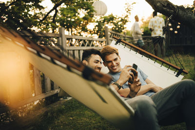 Cheerful friends using smart phone while resting in hammock at yard