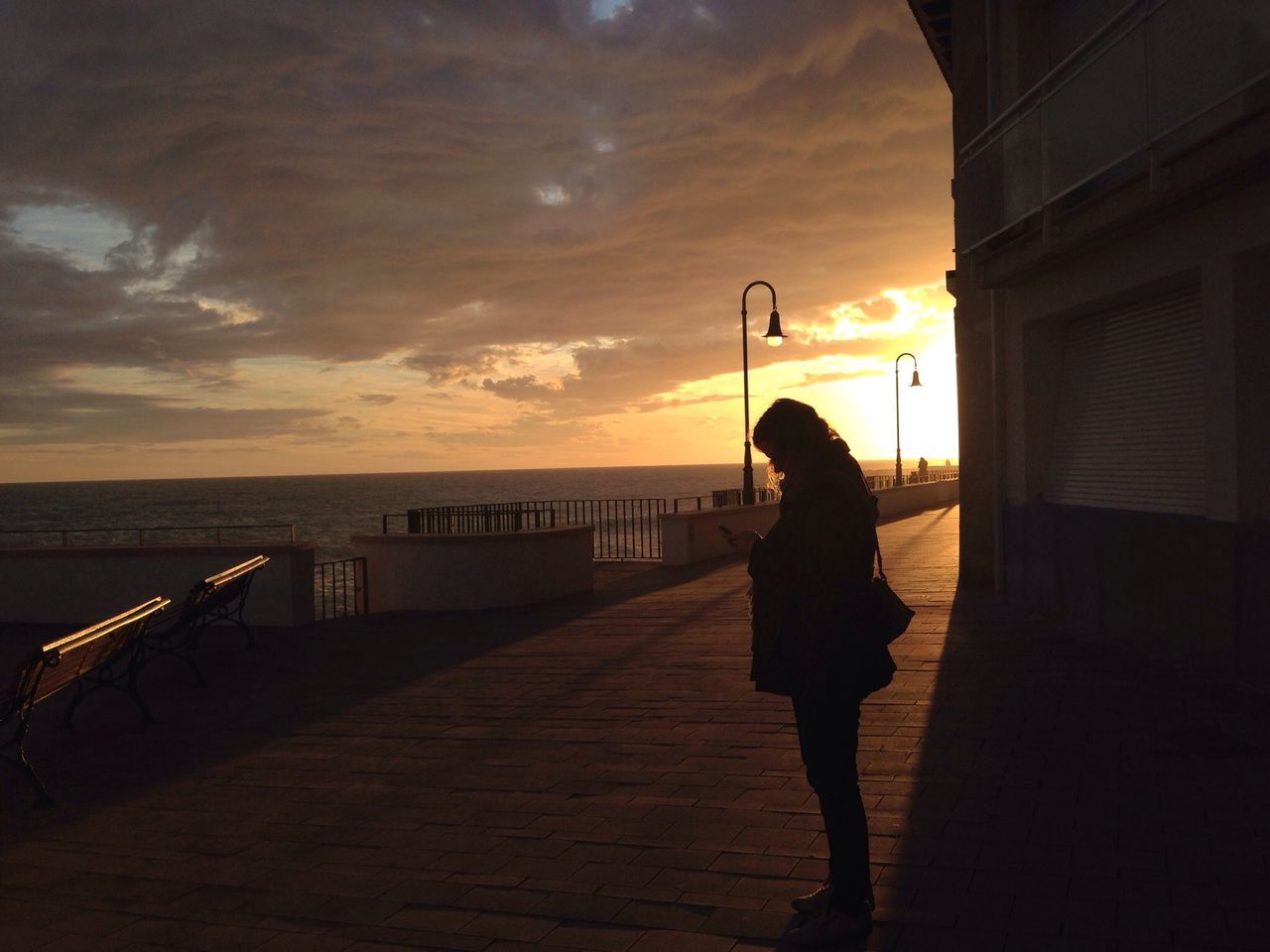 sunset, sky, lifestyles, standing, sea, full length, leisure activity, silhouette, railing, water, orange color, sun, built structure, cloud - sky, rear view, person, horizon over water, sunlight