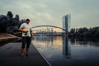 Portrait of man fishing while standing by river in city during sunset