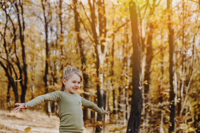 Smiling caucasian child girl playing with yellow leaves in autumn forest or garden around trees