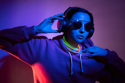 Cyberpunk woman in hooded hoodie and sunglasses dances against wall with neon sticks and headphones