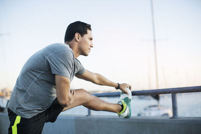 Thoughtful male athlete stretching leg on pier by harbor