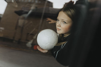 Contemplative girl with soccer ball seen through glass of car