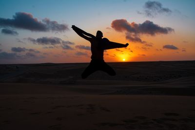 Silhouette man jumping on sand against sky during sunset