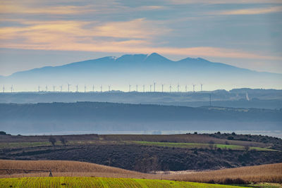 Layered scenic view of field against hill, wind turbines, cloud, mountain and sky