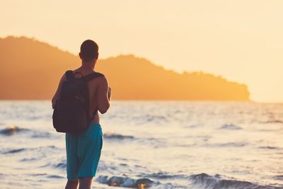 Rear view of man carrying backpack looking at sea during sunset