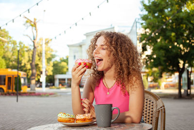 Young attractive curly woman sits at table in cafe on summer terrace. colorful delicious donuts,