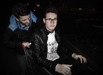 Young man pushing shopping cart while friend sitting on it at night