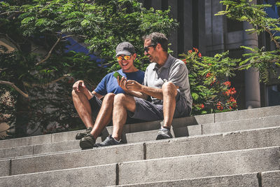 Father showing smart phone to son while sitting on steps in city during sunny day