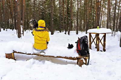 Young woman in yellow from behind sitting on snow covered bench in winter forest and holding dog 