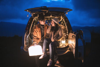 Young couple sitting in illuminated car trunk at night