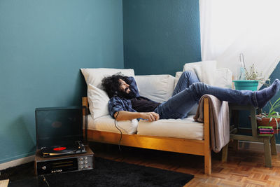 Bearded man listening music through turntable while lying on sofa at home