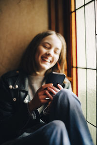 Smiling girl using smart phone while sitting by window at home