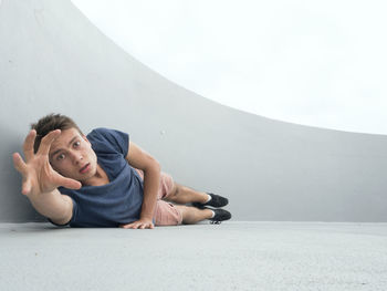 Portrait of young man gesturing while lying by wall on footpath