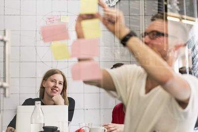 Young businessman writing ideas on adhesive notes with colleagues in background at creative office