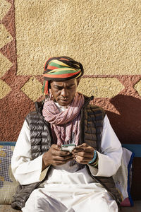 Serious egyptian male wearing traditional turban and clothes surfing internet on smartphone while sitting on sofa in courtyard near house on sunny day