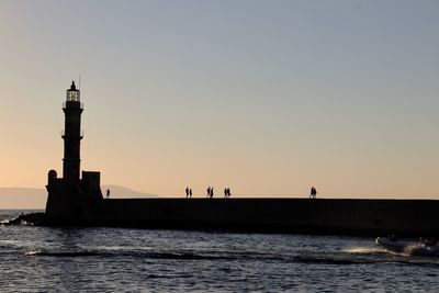Silhouette people walking on pier towards lighthouse during sunset