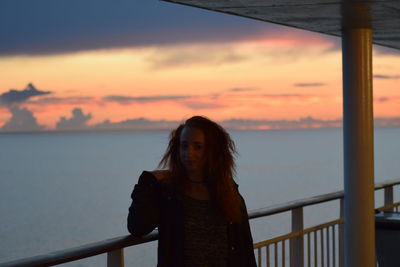 Portrait of young woman standing by railing against sea during sunset